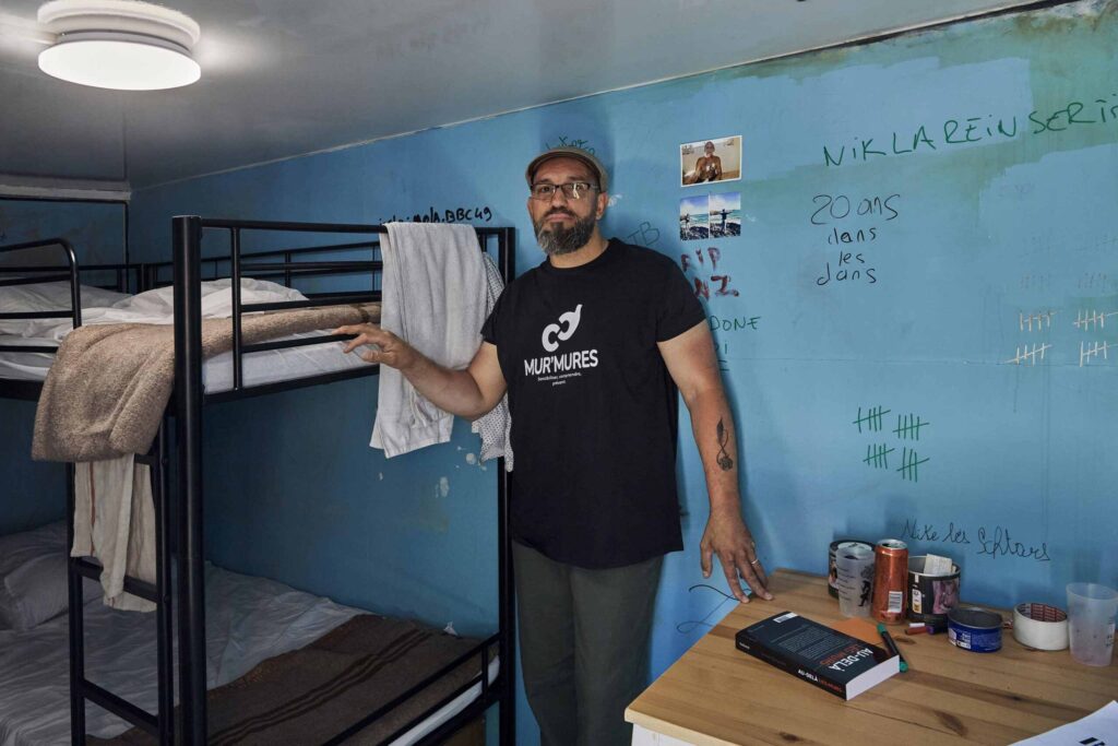 Yannick Deslandes dans sa cellule mobile, avec des lits superposés et des graffitis au mur.
